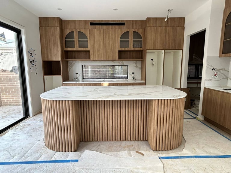 Modern kitchen under construction with curved wood-grain island, white countertops, and light wood cabinetry