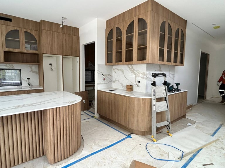 Modern kitchen under construction with wooden cabinetry, marble countertops, curved island, and appliances being installed