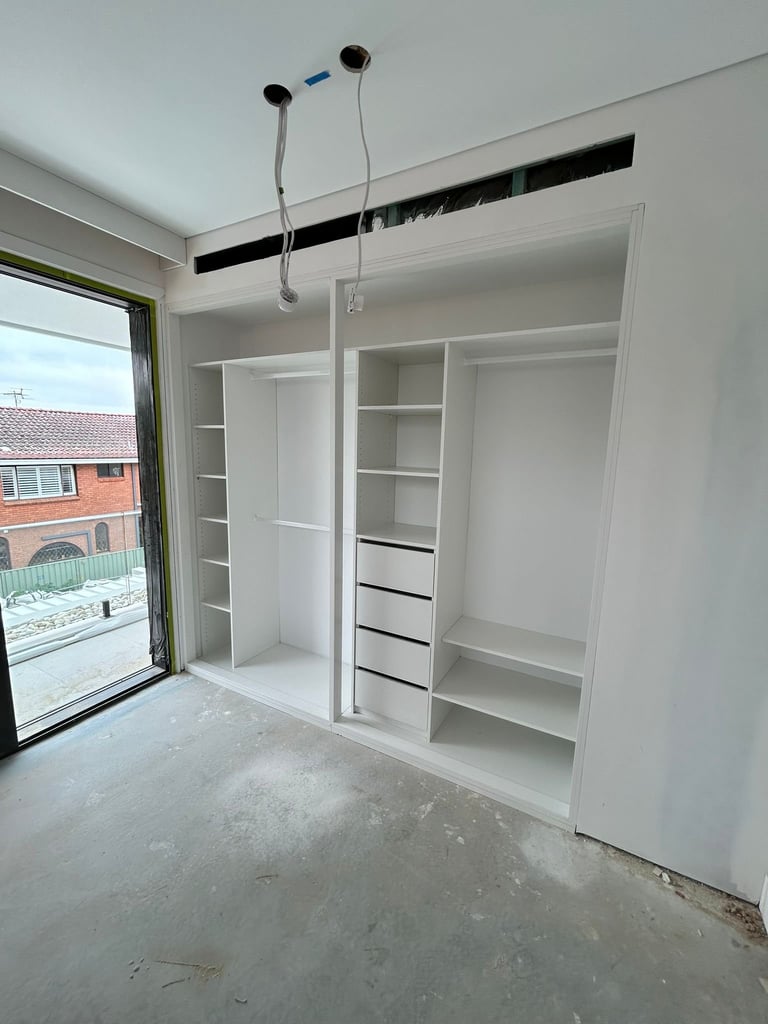 Modern walk-in closet under construction with white shelving units, drawers, pendant lighting fixtures, and glass door opening to courtyard
