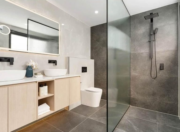 Modern bathroom with light wood vanity, white vessel sinks, large mirror, and walk-in shower with dark gray tile and glass enclosure