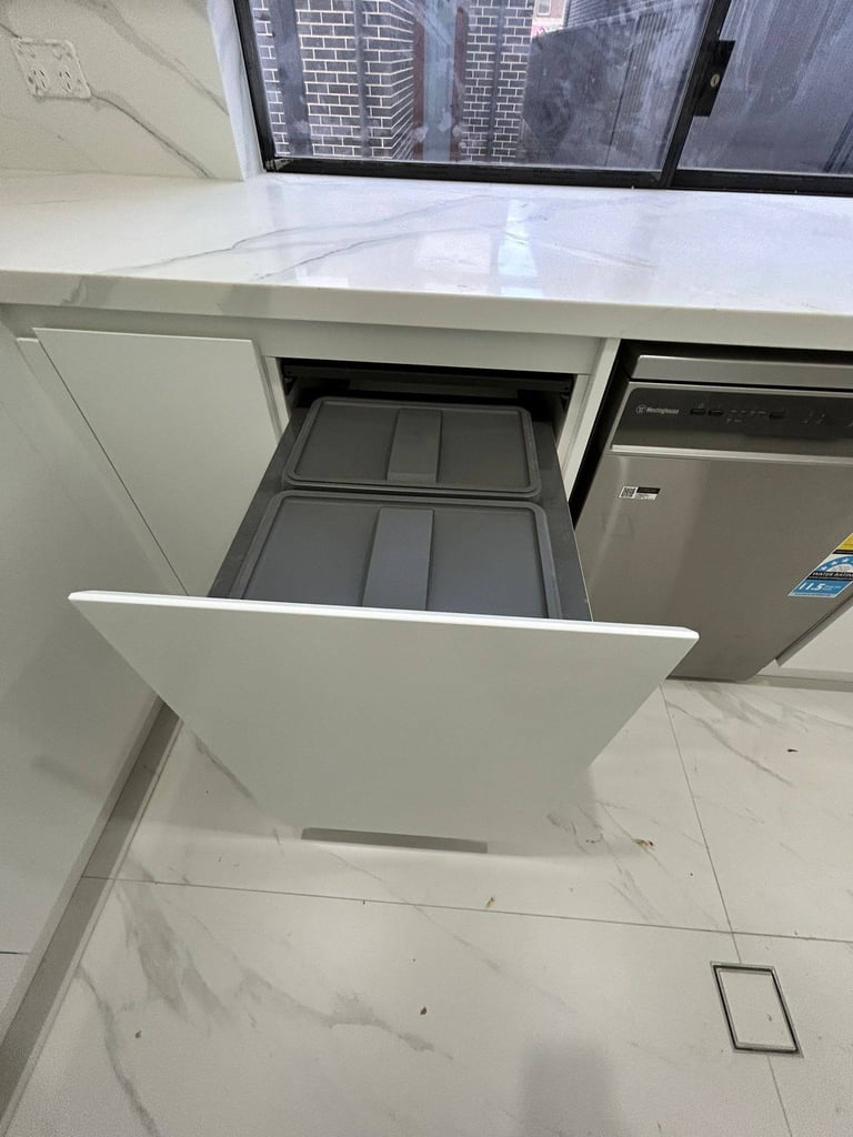 Modern kitchen with white cabinetry, open drawer showing gray compartments, stainless steel appliances, and city skyline visible through windows above.