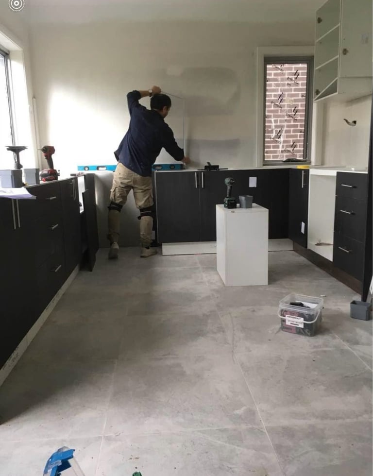 Person in navy jacket standing in unfinished kitchen with black cabinets and concrete floor during renovation