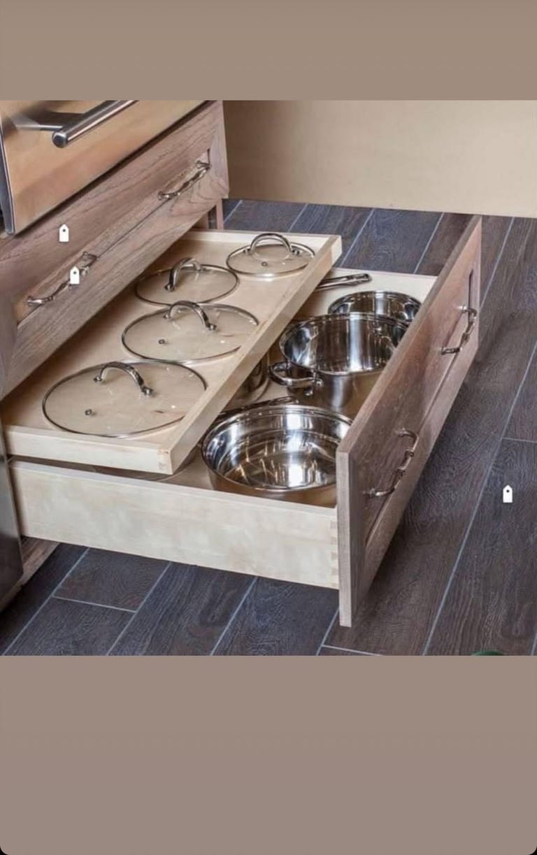 Open kitchen drawer organizer displaying pot lids on left and stainless steel cookware on right, inside modern wooden cabinet with gray tile flooring