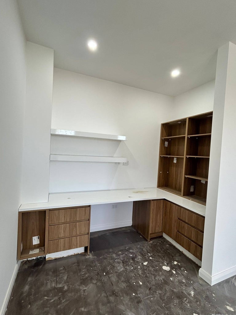 Modern home office with wood cabinetry, white countertop desk, floating shelves, and recessed ceiling lights on concrete flooring