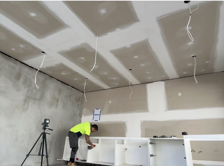 Worker in neon yellow vest installing drywall on ceiling with grid pattern and hanging wires in unfinished interior space