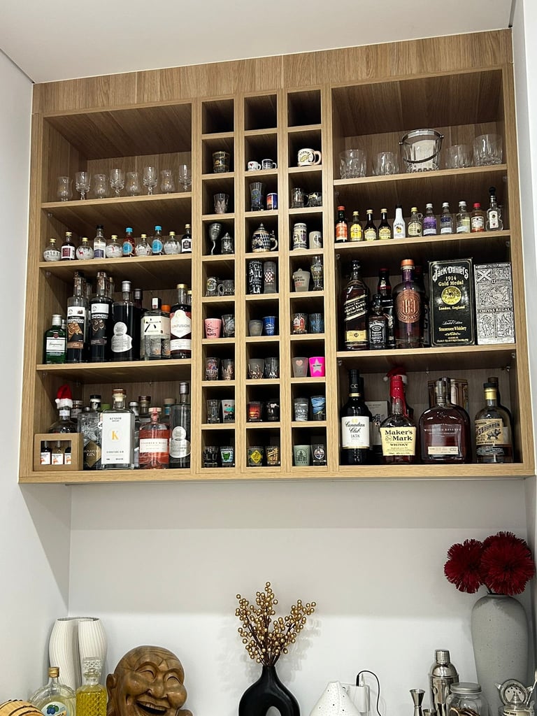Wooden wall-mounted liquor cabinet with multiple shelves displaying various spirits, bottles, and glassware, with decorative items below